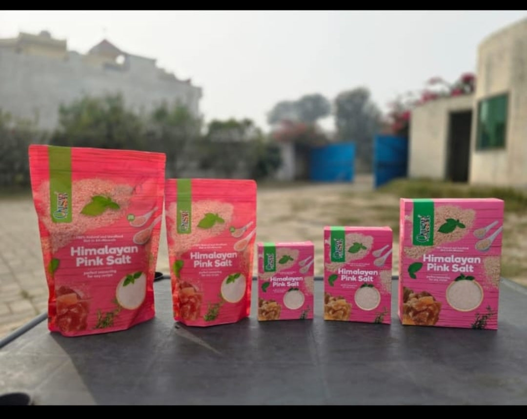 Four packages of Himalayan Pink Salt in various sizes arranged in a row on a dark table, with a blurred outdoor background of buildings and trees.